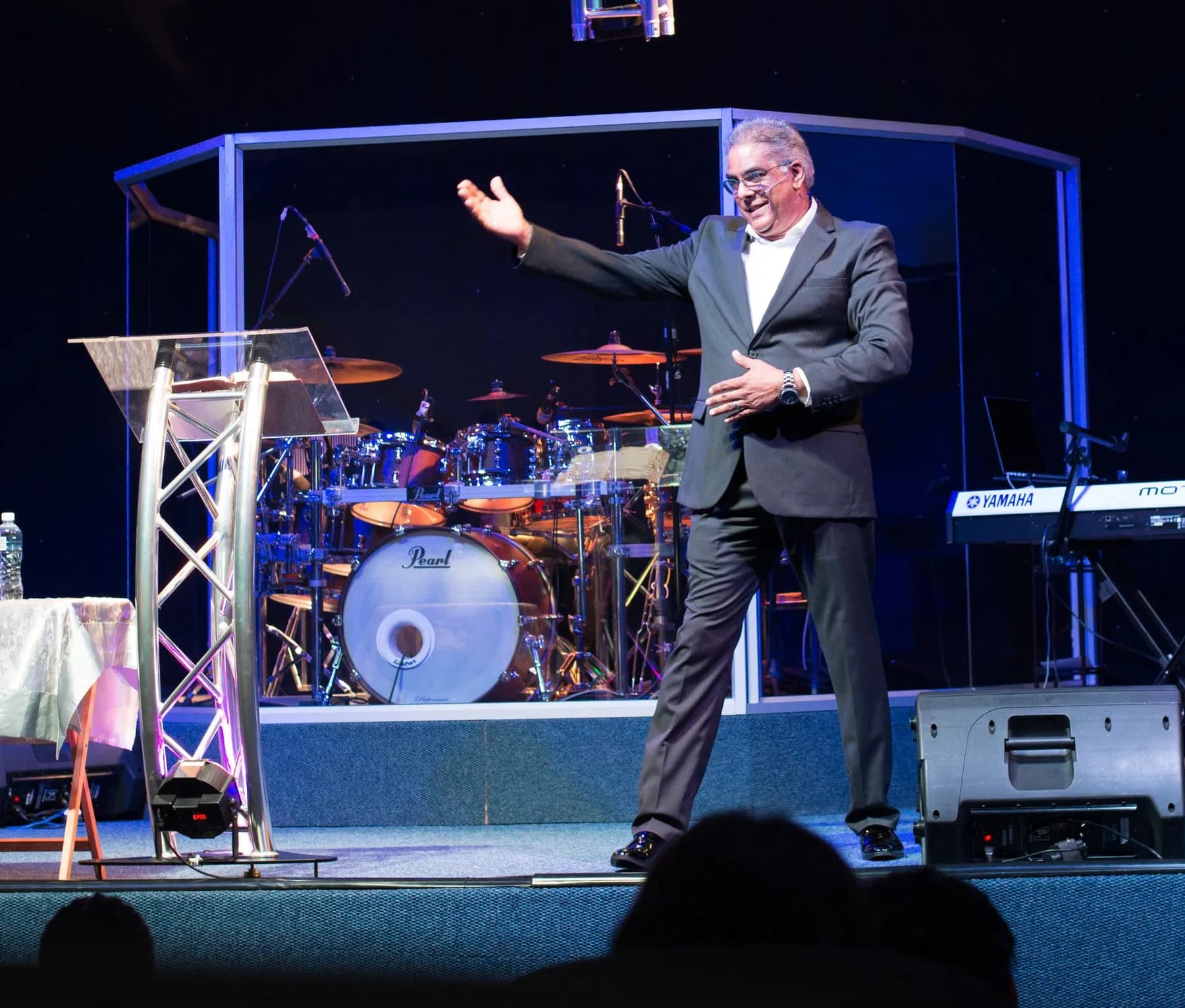 Dr RJ Sing on stage in a grey suit, arms open in welcome
